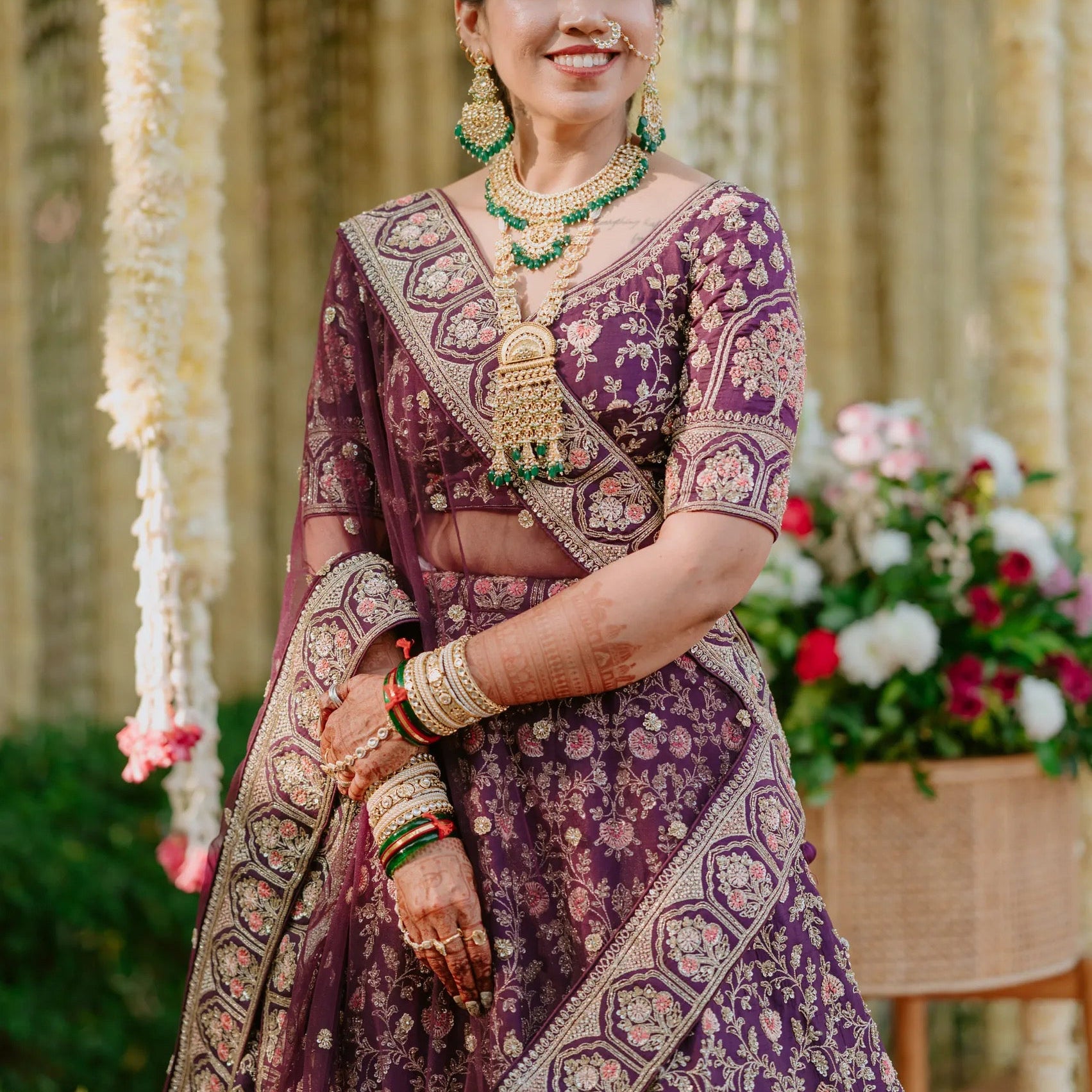 Woman in traditional purple and gold saree with jewelry, standing outdoors with floral decorations.