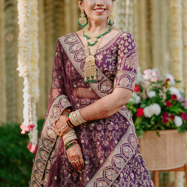 Woman in traditional purple and gold saree with jewelry, standing outdoors with floral decorations.