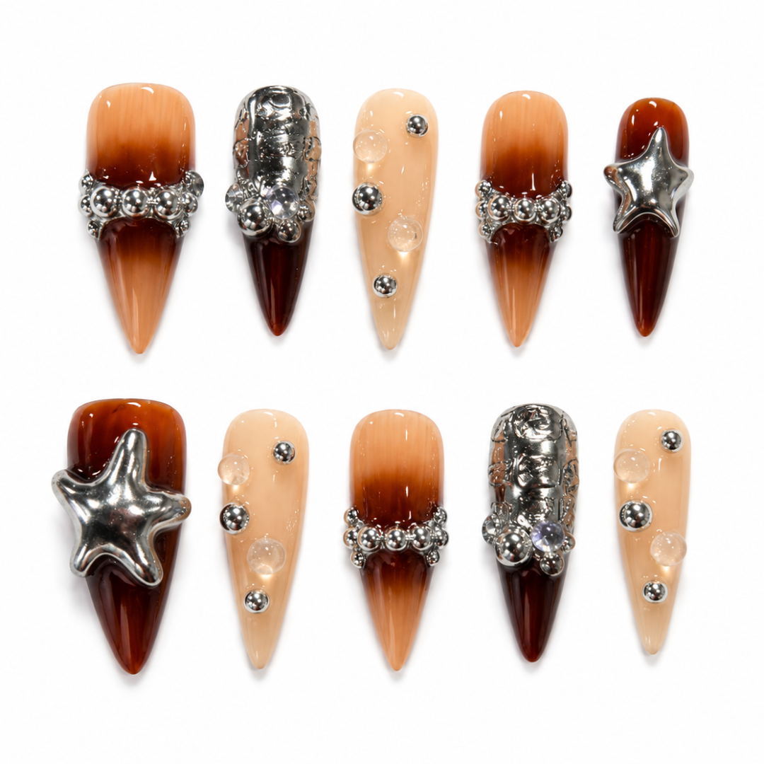 Twelve pointed artificial nails arranged in two rows on a white background. The set includes gradient designs in peach, orange, and deep brown, embellished with silver rhinestone bands, clear bubble accents, and metallic star charms.