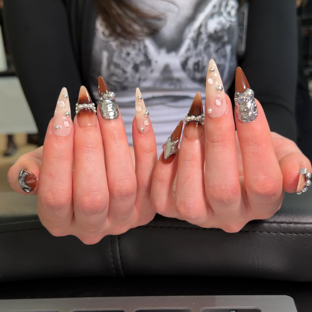 A close-up of a person's hands displaying a set of long, decorated press-on or nail art nails. The nails are primarily nude beige with silver glitter, rhinestones, and metallic silver designs.