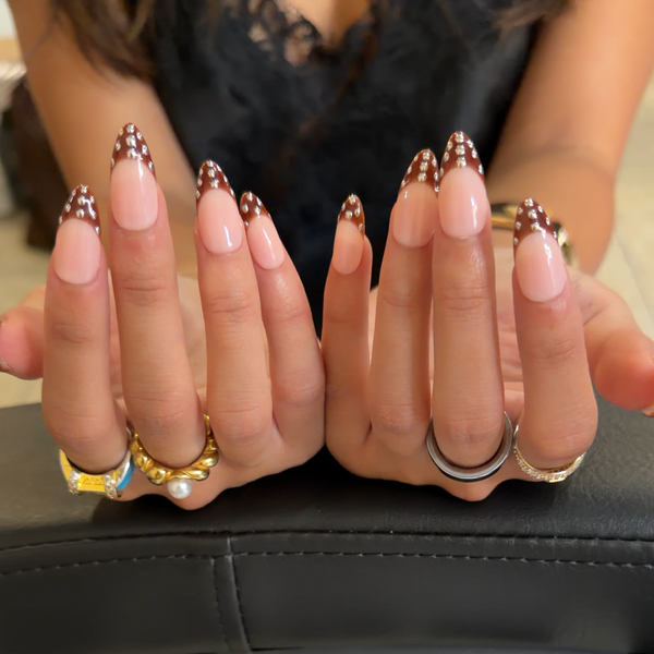 A close-up of a person's hands displaying a manicure with long, glossy pink nails and metallic gold rhinestone accents. The person is wearing multiple rings, and the background shows a black leather surface.