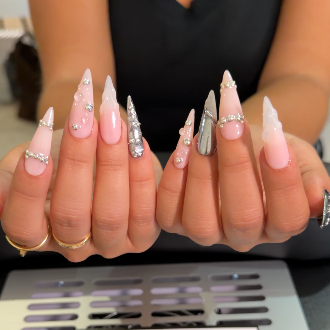 A close-up of a person's hands displaying long, sculpted press-on or nail art nails in a light pink and white gradient with rhinestone accents. A card reading 'OVALIS BLISS' is visible in the foreground.