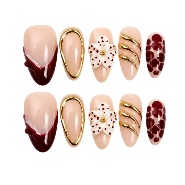 Six-piece press-on nail set arranged in two rows of three on a plain white background. The nails have a glossy nude-peach base with decorative designs including dark burgundy French tips, gold metallic swirls, a white flower with red dots, and a burgundy polka-dot pattern.