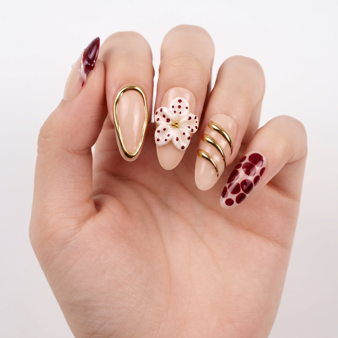 A close-up of a hand showcasing a variety of nail art designs and press-on nails. The nails feature different styles: a dark burgundy oval, a white flower with red dots and a gold center, three gold curved bands, a burgundy flower with red dots, and a solid burgundy oval.