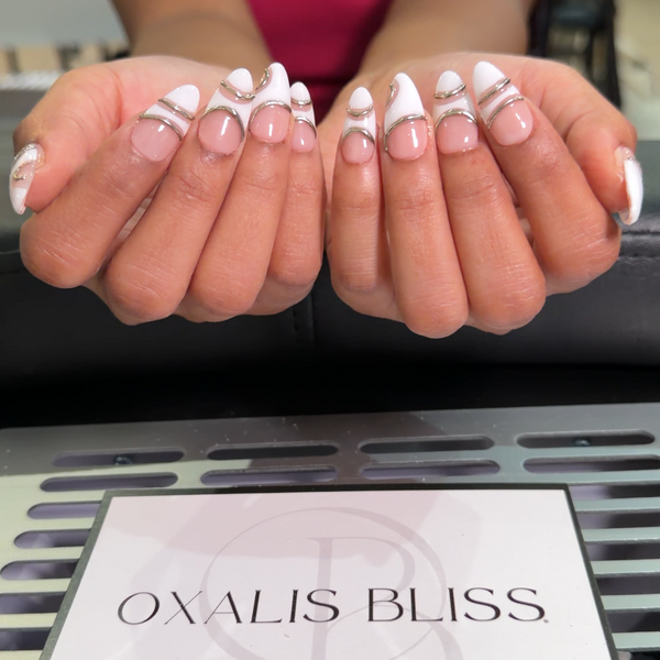 A close-up of a person's hands displaying a white and clear French manicure with decorative nail art, with a salon sign reading 'OXALIS BLISS' in the foreground.